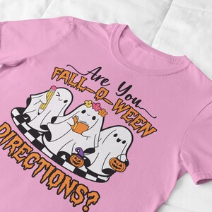 K&ouml;nnte beinhalten: Rosafarbenes T-Shirt mit einem Halloween-Motiv. Das Design zeigt drei Cartoon-Geister mit Bleistift, Buch und Blumenkranz sowie K&uuml;rbissen. Der Text lautet "Are You Fall-O-Ween Directions?" in Orange und Schwarz.