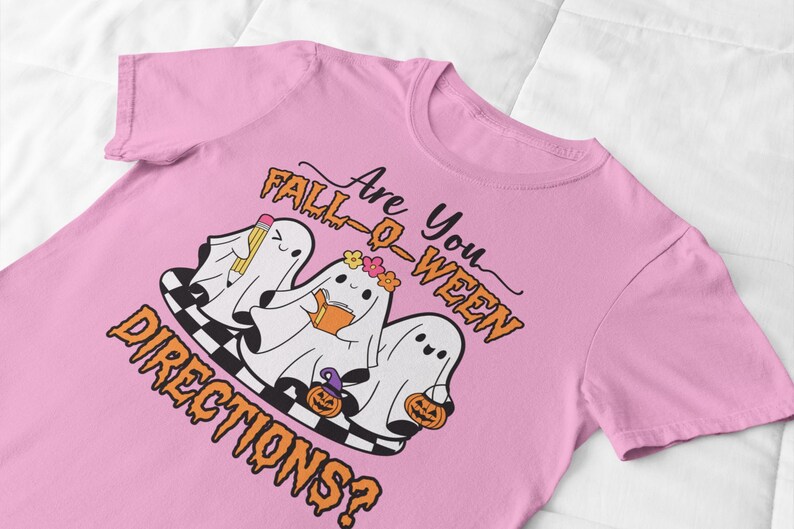 K&ouml;nnte beinhalten: Rosafarbenes T-Shirt mit einem Halloween-Motiv. Das Design zeigt drei Cartoon-Geister mit Bleistift, Buch und Blumenkranz sowie K&uuml;rbissen. Der Text lautet "Are You Fall-O-Ween Directions?" in Orange und Schwarz.