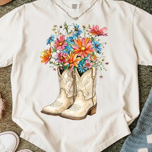 May include: Cream-coloured t-shirt featuring a colourful floral bouquet spilling out of a pair of cream-coloured cowboy boots. The flowers are in shades of pink, orange, blue, and yellow. The boots have decorative stitching.
