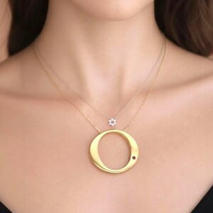 May include: A gold-colored necklace with a large letter "O" pendant and a small red gemstone. A second necklace features a small, six-pointed star pendant. The necklaces are worn on a person's neck.