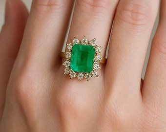 May include: A gold ring featuring a large, rectangular, emerald-cut green gemstone at the center, surrounded by a halo of small, round, clear white diamonds set in a scalloped, pronged arrangement on a thin, polished gold band.