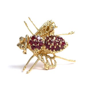 May include: A gold-colored insect brooch with ruby-colored gemstones. The brooch features detailed wings, legs, and antennae. The body of the insect is covered in small, round, ruby-colored stones. The brooch is set against a white background.
