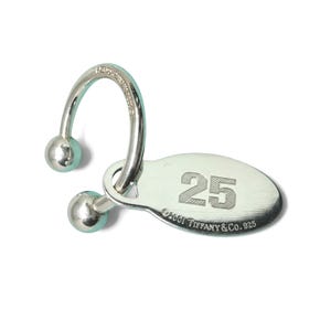 May include: A silver-toned metal keychain featuring a curved bar with two spherical ends and an attached oval tag. The tag is engraved with the number 25 and the text ©2001 Tiffany & Co. 925. The curved bar also features small engraved text.