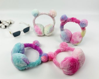 May include: Three pairs of colorful fuzzy earmuffs with pom poms. The earmuffs are in shades of pink, blue, and purple.