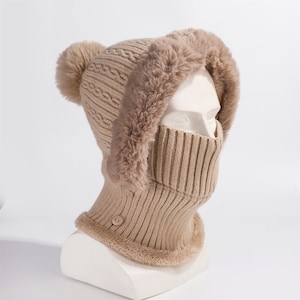 Peut inclure: Un bonnet cagoule en tricot beige avec une bordure en fausse fourrure autour du capuchon et du visage. Le chapeau présente un motif en tricot torsadé sur le dessus et un pompon assorti. Le protège-cou a deux boutons. Ce bonnet d'hiver est conçu pour vous garder au chaud.