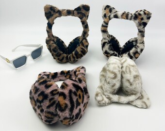 May include: Four pairs of leopard print earmuffs with cat ear details. The earmuffs are in shades of brown, black, pink, and white. A pair of white sunglasses with black lenses are in the background.