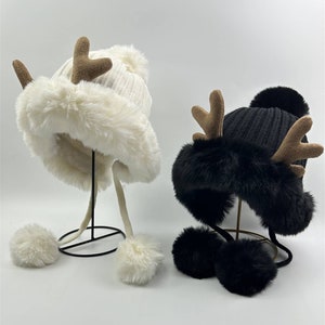 May include: Two winter hats, one white and one black, each with faux fur trim and brown antler details. The white hat has a matching pom-pom and ear flaps with pom-pom ends. The black hat has a black pom-pom and ear flaps.