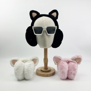 May include: A black faux fur earmuff headband with cat ears and gold glitter accents, worn by a mannequin head with white sunglasses. Two matching earmuffs, one white and one pink, are also displayed.