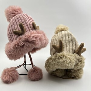 May include: Two knitted winter hats with faux fur trim and pom poms. The hats have reindeer antlers on top. One hat is pink and the other is beige.