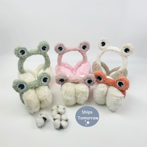 May include: Four plush ear warmers shaped like frogs in green, pink, brown, and orange. Each ear warmer has large white eyes and a white fuzzy interior.