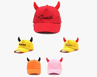 May include: Five different colored baseball caps with devil horns. The caps are red, yellow, orange, pink, and yellow. The text on the caps reads "Smooth", "Custom", "Bandakhela", and "Custom".