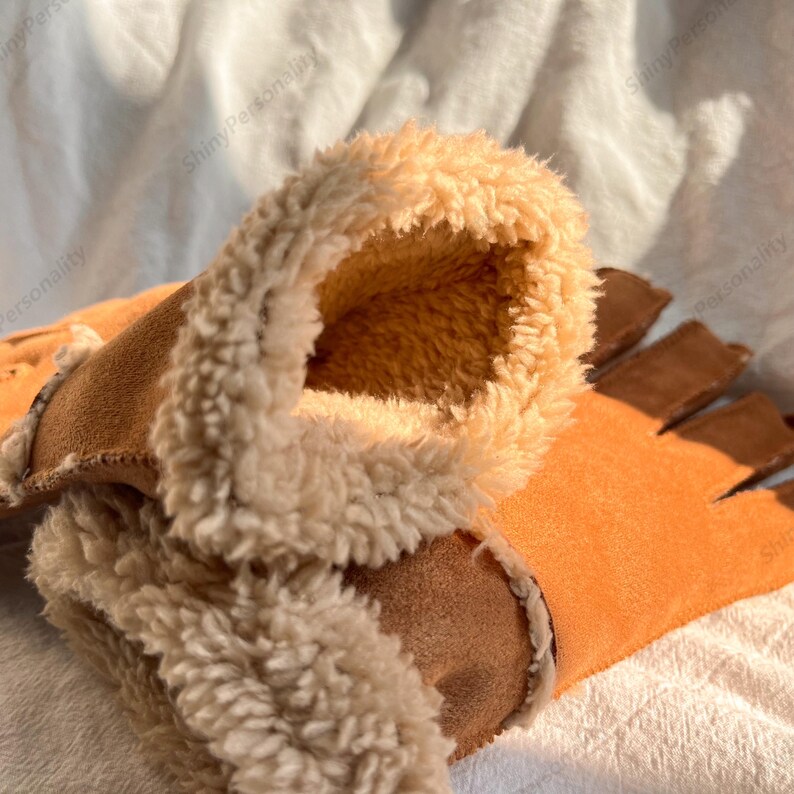 May include: Close-up of a pair of fingerless gloves. The gloves are a warm orange color with a brown suede-like material on the outside and a soft, cream-colored faux fur lining the inside. The gloves are designed to keep hands warm while leaving fingers free.