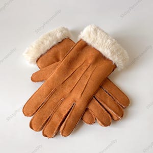 May include: A pair of brown suede gloves with a cream-colored faux fur cuff. The gloves are lying on a white surface. The gloves have a classic design and are likely for cold weather.