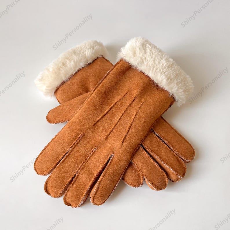 May include: A pair of brown suede gloves with a cream-colored faux fur cuff. The gloves are lying on a white surface. The gloves have a classic design and are likely for cold weather.