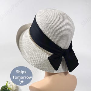May include: A white straw hat with a black ribbon and pearl accents. The hat has a rounded crown and a wide brim. A black bow is tied on the side. The text "Ships Tomorrow" is on the bottom left.
