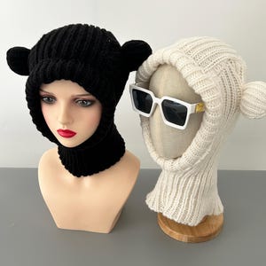 Könnte beinhalten: Zwei Strickmützen mit Ohrenklappen in Form von Bärenohren. Eine Mütze ist schwarz und die andere ist weiß.