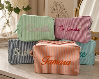 May include: Five rectangular fabric pouches with vertical stripes in light green, pink, dark blue, orange, and teal. Each pouch features embroidered names including Camille, Lia Amada, Sutton, Tamara, and a partial name, with a small daisy on one.