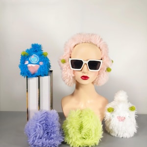 Pode incluir: Quatro brinquedos de pelúcia com texturas felpudas. Um é um chapéu de pele rosa com óculos de sol brancos. Outro é um monstro azul e rosa com detalhes verdes. O terceiro é um monstro branco com detalhes verdes. O último é uma bola felpuda de cor roxa clara.