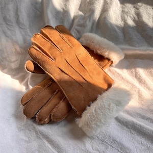May include: A pair of tan suede gloves with white faux fur cuffs. The gloves are lying on a white surface. The gloves have visible stitching and a soft texture, suitable for cold weather.