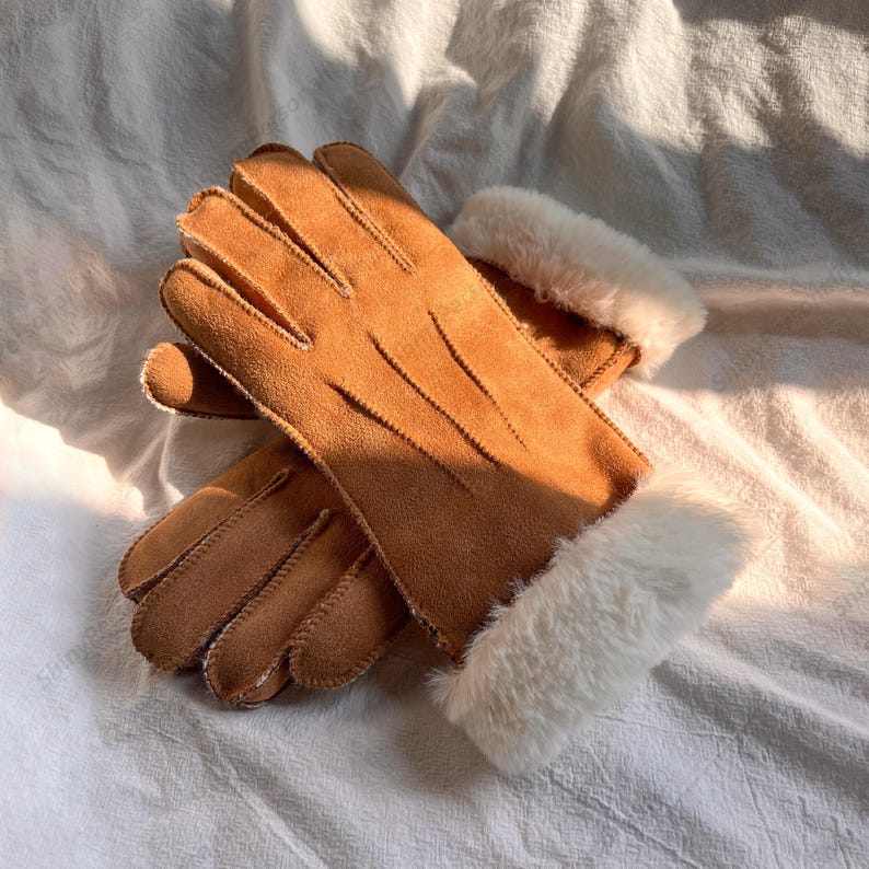 May include: A pair of tan suede gloves with white faux fur cuffs. The gloves are lying on a white surface. The gloves have visible stitching and a soft texture, suitable for cold weather.