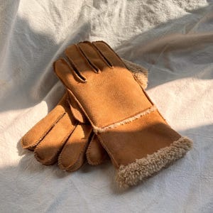 May include: A pair of tan gloves made of suede-like material with a soft, fuzzy lining. The gloves have a classic design with individual fingers and a warm, cozy appearance, perfect for cold weather.