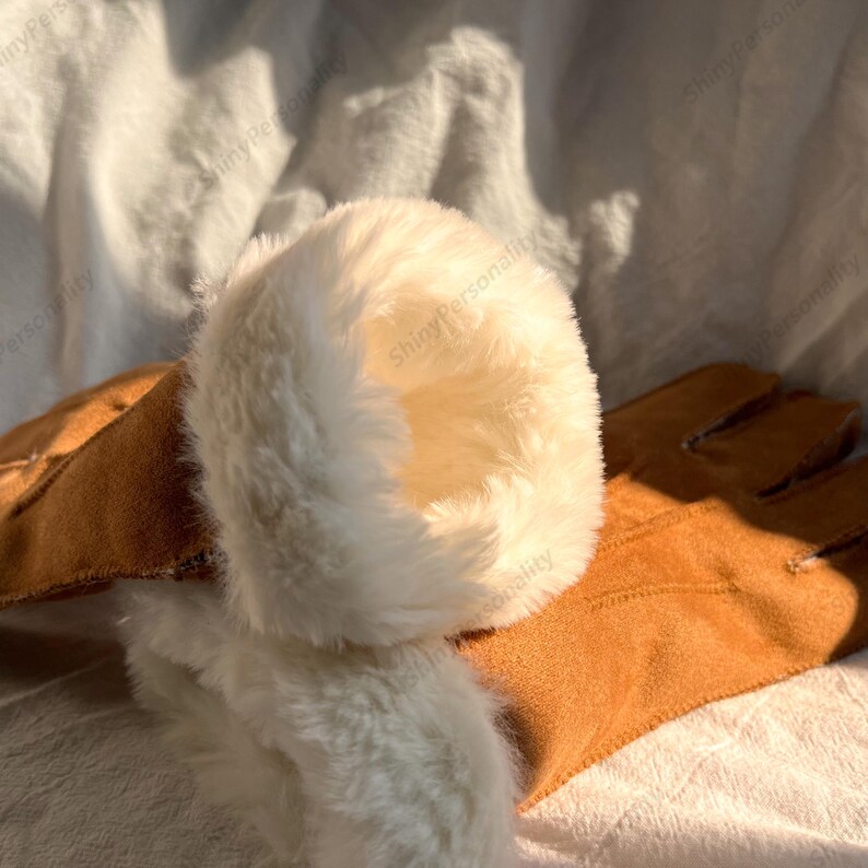 May include: A pair of brown gloves with a soft, white, faux fur lining around the wrist. The gloves are made of a suede-like material and appear warm and cozy. The gloves are resting on a white surface.