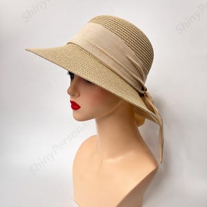 May include: A wide-brimmed straw hat with a beige ribbon tied around the crown. The hat is on a white mannequin head.
