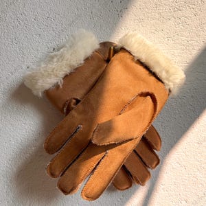 May include: A pair of brown gloves with a soft, suede-like texture. The gloves have a cream-colored, fluffy trim around the wrists. The gloves are layered on top of each other, showing the stitching details.