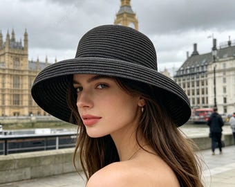 May include: A black sun hat with a wide brim. The hat is made of a woven material and has a rounded crown. The background features a cloudy sky and a historic building with a clock tower.
