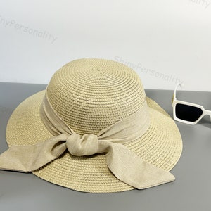 May include: A beige straw hat with a wide brim and a large bow tied at the front. The hat is made of woven straw and has a natural, rustic look.