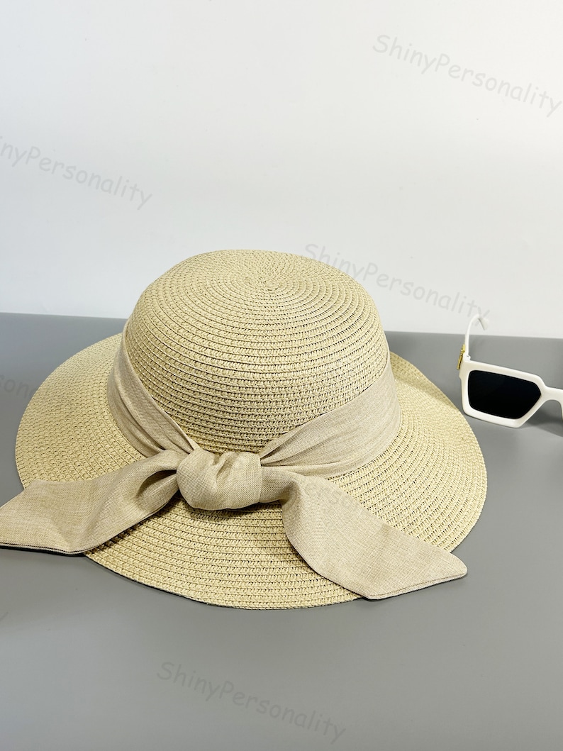 May include: A beige straw hat with a wide brim and a large bow tied at the front. The hat is made of woven straw and has a natural, rustic look.