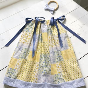 May include: A sleeveless dress featuring a patchwork pattern of yellow gingham, yellow floral, and blue botanical prints, finished with a solid light blue hem and dark blue ribbon shoulder ties, accompanied by a matching light blue bow headband.