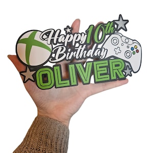 Pode incluir: Um topo de bolo de aniversário com tema de jogos. Apresenta um logotipo Xbox verde e branco, um controle branco e as palavras "Happy 10th Birthday OLIVER" em verde e branco. O design inclui estrelas.