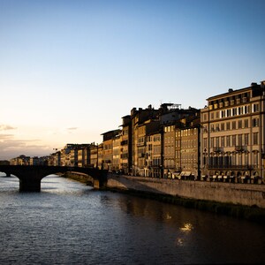 May include: A view of a bridge over a river in a city. The river is calm and the water is reflecting the light of the setting sun. Buildings line the riverbank on the right side of the image.