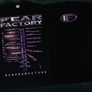 Puede incluir: Camiseta negra con un gráfico morado y blanco de un esqueleto con el texto "FEAR FACTORY" y "DEMANUFACTURE". La parte posterior de la camiseta tiene una "F" morada en un círculo.