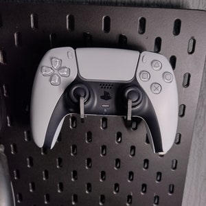 May include: A white PlayStation 5 DualSense wireless controller with black buttons and a black PlayStation logo. The controller is hanging on a black pegboard.