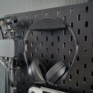 May include: Black over-ear headphones hanging on a black pegboard. The headphones have black earcups and a silver band. A webcam and a handheld gaming device are also visible.