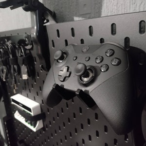 May include: A black Xbox controller with a textured surface and black buttons. The controller is mounted on a black pegboard wall.