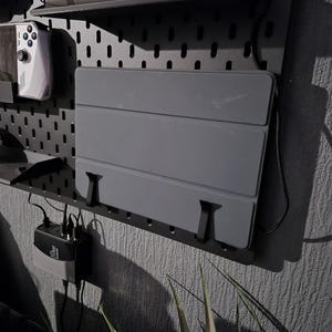 May include: A grey tablet case is mounted on a black pegboard wall organiser. The case is held in place by black brackets. A gaming console and a computer mouse are on a shelf above the case. A power adapter is plugged into an outlet below.