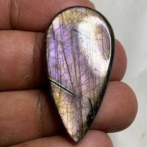 May include: A teardrop-shaped labradorite gemstone with a polished surface. The stone displays a mix of purple, gold, and black colors with visible internal fractures. The gemstone is held in a hand.