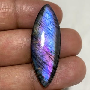 Può includere: Una gemma allungata e lucidata, che mostra un gioco di colori. La pietra presenta un mix di tonalità viola, blu e iridescenti. La gemma misura circa 3,8 cm di lunghezza e ha una superficie liscia e riflettente.