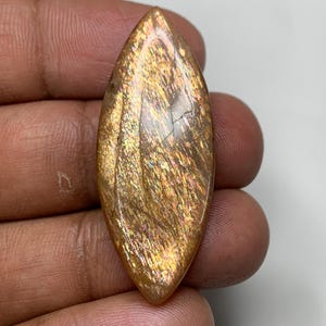 May include: A polished, oval gemstone in warm golden-orange tones. The stone has a shimmering, iridescent effect with streaks of light and colour. The gemstone is held in a hand, showing its size and shape.