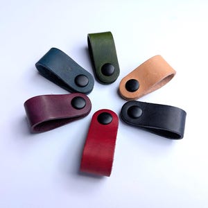 May include: Six leather cable tidies in a range of colours: navy, green, tan, black, burgundy, and red. Each tidy has a loop design with a black snap fastener. The tidies are arranged in a circle against a white background. Each is designed to keep cables neat.