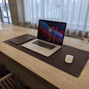 May include: A silver laptop and a tablet are on a brown leather desk mat. A white mouse is to the right of the laptop. The laptop screen shows a colourful graphic. The desk is made of light wood.
