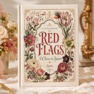 May include: A hardcover book with a cream-colored cover featuring a decorative floral border in shades of pink, red, and green. The text on the cover reads A LITTLE BOOK OF RED FLAGS I Chose to Ignore Again in elegant red and black lettering.