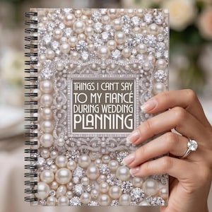 May include: A spiral-bound notebook with a cover adorned with faux pearls and rhinestones. The notebook features the text "THINGS I CAN'T SAY TO MY FIANCÉ DURING WEDDING PLANNING" in a decorative frame. The notebook is held by a person.