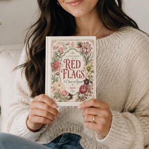 May include: A hardcover book titled A Little Book of Red Flags I Chose to Ignore Again. The cover is cream-colored with a decorative gold border and a vibrant floral illustration featuring pink, red, and green flowers surrounding the red text.