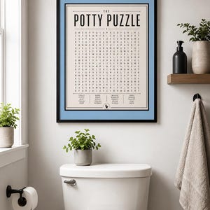 K&ouml;nnte beinhalten: Ein gerahmter Wandkunstdruck mit dem Titel The Potty Puzzle, der ein Wortsuchr&auml;tsel mit schwarzem Text auf cremefarbenem Hintergrund zeigt, eingefasst in ein hellblaues Passepartout und einen d&uuml;nnen schwarzen Rahmen mit Wortliste und WC-Symbol.