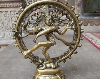 May include: A brass-colored metal statue of a dancing figure with multiple arms, positioned within a circular flame-like ring. The figure stands on a lotus-shaped base. The text "STYLE - 1" is visible at the top, and "LIVKASHI" is at the bottom.