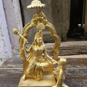 May include: A detailed brass sculpture of a deity seated on a throne, holding a mace. The sculpture is golden in color and features intricate details, including a decorative arch and a smaller figure kneeling nearby. The statue is on a rectangular base.